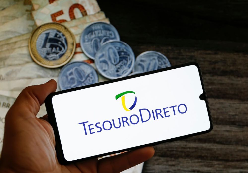 Tesouro direto 2025. Celular mostrando site do tesouro direto e com algumas notas de dinheiro e moedas atrás