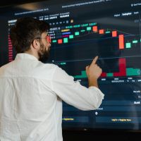 Homem de camisa branca apontando uma tela onde possui um gráfico de ações da bolsa de valores